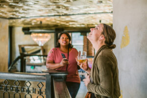 Two women touring a stylish Colorado venue; one smiles while looking at the gold ceiling with a drink in hand, the other listens attentively with phone in hand—highlighting the guest experience and planning process.