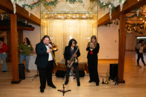 Three jazz musicians performing indoors at a Boulder event venue — playing trombone, saxophone, and trumpet beneath greenery and string lights — representing live music and event entertainment in Colorado.